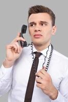 trött av kontor arbete. deprimerad ung man i skjorta och slips talande på de telefon och innehav telefon tråd medan stående mot grå bakgrund foto