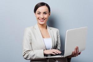 stödjande din företag. självsäker ung affärskvinna innehav bärbar dator och leende medan stående mot grå bakgrund foto