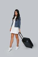 redo för resa. full längd av skön ung kvinna leende och dragande bagage medan stående mot grå bakgrund foto