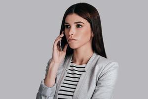företag prata. attraktiv ung kvinna i smart tillfällig ha på sig talande på de telefon och ser bort med allvarlig ansikte medan stående mot grå bakgrund foto