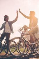 hög fem till vän. låg vinkel se av glad ung människor stående nära deras cyklar på de väg medan två män ger hög fem till varje Övrig foto