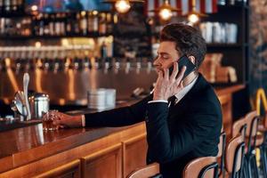 stilig ung man i full kostym talande på smart telefon medan Sammanträde på de bar disken i restaurang foto