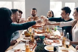 vem vill ha Mer pizza grupp av ung människor i tillfällig ha på sig plockning pizza och leende medan har en middag fest inomhus foto