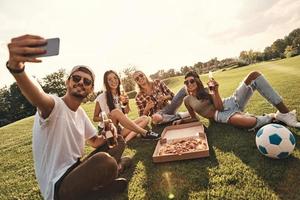 gående galen tillsammans. grupp av ung leende människor i tillfällig ha på sig tar selfie medan njuter mat och drycker utomhus foto