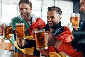 Lycklig ung män täckt i internationell flaggor toasting med öl medan tittar på sport spel i de pub foto