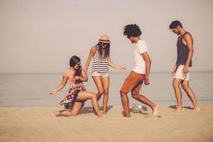 strand boll med vänner. grupp av glad ung människor spelar med fotboll boll på de strand med hav i de bakgrund foto