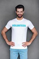 herr. volontär. självsäker ung man i volontär- t-shirt förvaring händer på höfter och ser på kamera med leende medan stående mot grå bakgrund foto