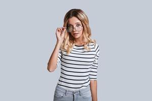 känsla nyfiken. attraktiv ung kvinna justeras henne glasögon och ser på kamera medan stående mot grå bakgrund foto