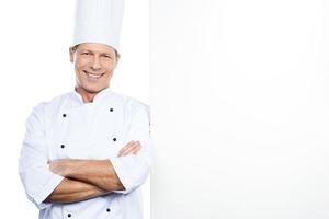 glad kock. självsäker mogna kock i vit enhetlig lutande på de kopia Plats och leende medan stående mot vit bakgrund foto