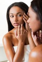 är den acne koncentrerad ung afrikansk kvinna rörande henne ansikte och ser på själv i en spegel foto