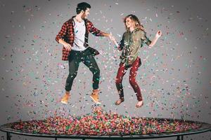 färgrik roligt. i luften skott av skön ung glad par innehav händer medan Hoppar på trampolin tillsammans med konfetti Allt runt om dem foto