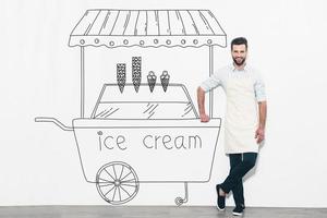 drömma handla om ny företag. stilig ung man i förkläde stående i främre av de vägg och lutande på de penna dragen is grädde vagn foto
