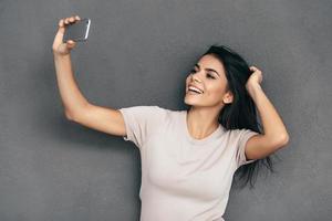 selfie tid attraktiv ung kvinna i tillfällig ha på sig framställning selfie förbi henne smart telefon och leende medan stående mot grå bakgrund foto