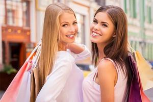 njuter handla tillsammans. bak- se av två skön ung kvinnor innehav handla påsar och ser över axel med leende medan stående utomhus foto