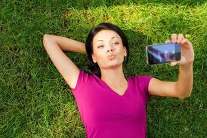 kvinna framställning selfie. topp se av skön ung kvinna framställning selfie förbi henne smart telefon medan liggande i grön gräs foto