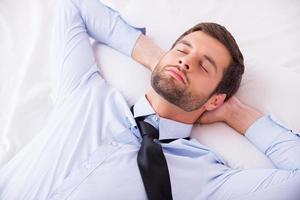 affärsman sovande. topp se av stilig ung man i skjorta och slips innehav händer Bakom huvud medan sovande i säng foto