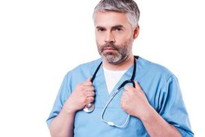 självsäker kirurg. självsäker mogna läkare i blå enhetlig ser på kamera och justeras hans stetoskop medan stående isolerat på vit foto