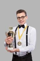 jag am vinnare glad ung man i rosett slips innehav en trofén och leende medan stående mot grå bakgrund foto