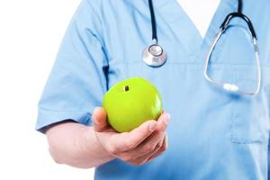 frukt göra du friska. närbild av kirurg i blå enhetlig innehav grön äpple medan stående isolerat på vit foto