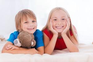 söt syster och bror. två söt barn ser på kamera och leende medan liggande tillsammans på säng foto