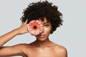 skön som blomma. attraktiv ung afrikansk kvinna ser på kamera och beläggning öga med en blomma medan stående mot grå bakgrund foto