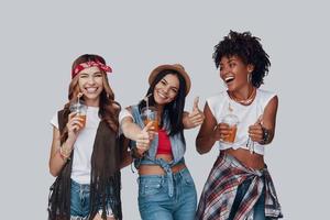 tre attraktiv ung kvinnor förvaring tummen upp och leende medan stående mot grå bakgrund foto