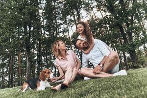 familj bindning tid. Lycklig ung familj av tre med hund leende medan Sammanträde på gräs i parkera foto