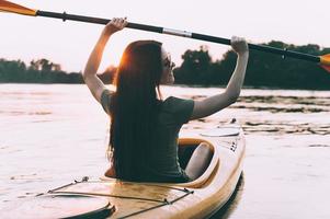 känsla Lycklig på de flod. bak- se av Lycklig ung kvinna Kajakpaddling på flod och höjning upp henne paddla med solnedgång i de bakgrund foto