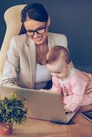 jag kan hjälp du topp se av liten bebis flicka använder sig av bärbar dator medan Sammanträde på kontor skrivbord med henne mor i kontor foto