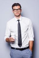 tar tid för kaffe ha sönder. självsäker ung man i skjorta och slips innehav kaffe kopp och ser på kamera medan stående mot grå bakgrund foto
