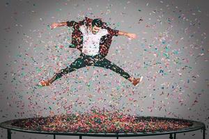 roligt i rörelse. i luften skott av stilig ung man Hoppar på trampolin med konfetti Allt runt om honom foto