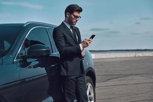 porträtt av förtroende. stilig ung affärsman använder sig av smart telefon medan stående nära hans bil utomhus foto