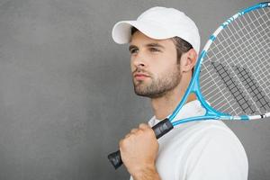 tennis är hans passion. självsäker ung man i sporter kläder bärande tennis racket på hans axel och ser bort medan stående mot grå bakgrund foto