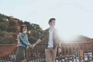 njuter trevlig dag tillsammans. skön ung par innehav händer och leende medan stående på de bro utomhus foto