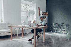 känsla släppte. stilig ung man kasta papper i luft och leende medan Sammanträde på hans arbetssätt plats i kreativ kontor foto