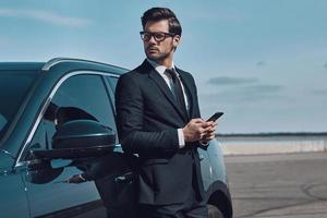 företag är hans liv. stilig ung affärsman använder sig av smart telefon medan stående nära hans bil utomhus foto