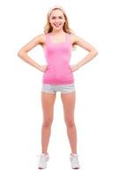sportig skönhet. full längd av skön utvikningsbrud blond hår kvinna i rosa skjorta innehav händer på höft och leende medan stående isolerat på vit bakgrund foto