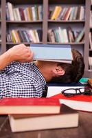 trött studerande. sida se av ung man beläggning hans ansikte med bok medan liggande på de hårt träslag golv med Övrig böcker om Allt runt om honom foto
