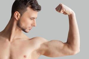 perfekt biceps. stilig ung muskulös man ser på hans biceps medan stående isolerat på grå bakgrund foto