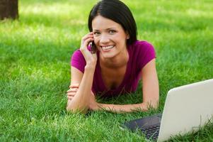 teknik framställning liv lättare. attraktiv ung kvinna talande på de mobil telefon och leende medan liggande i gräs med bärbar dator om nära henne foto