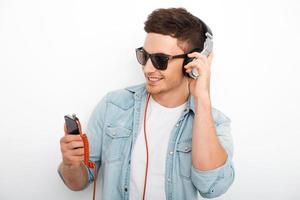 sorglös musik älskare. glad ung man i hörlurar leende och ser på smart telefon medan stående mot vit bakgrund foto