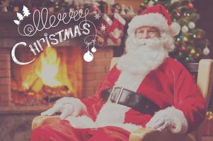 glad jul traditionell santa claus Sammanträde på hans stol och leende med öppen spis och jul träd i de bakgrund foto