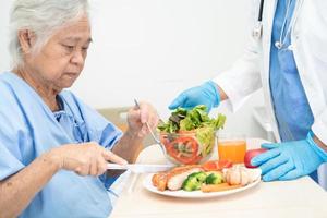 asiatisk senior eller äldre gammal dam kvinna patienten äter frukost och grönsaker hälsosam mat med hopp och glad medan du sitter och hungrig på sängen på sjukhuset. foto