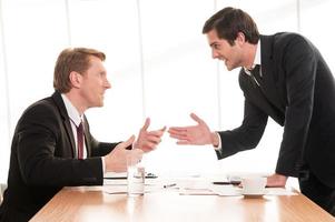 företag konflikt. två ung män i formell klädsel argumenterar och gestikulerar medan Sammanträde på de tabell foto