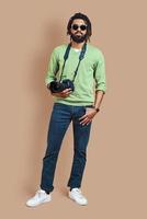 full längd av ung afrikansk fotograf i tillfällig Kläder ser på kamera medan stående mot brun bakgrund foto