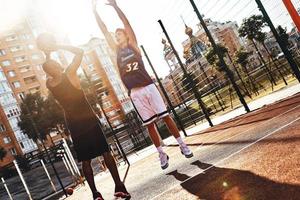 skytte de ring. två ung män i sporter Kläder spelar basketboll medan utgifterna tid utomhus foto