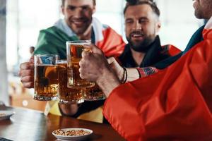 Lycklig ung män täckt i internationell flaggor toasting med öl medan tittar på sport spel i de pub foto