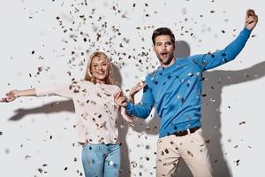 Semester är här skön ung par gestikulerar och leende medan stående mot grå bakgrund med konfetti flygande överallt foto