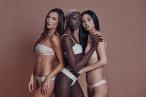 ser perfekt. tre attraktiv blandad lopp kvinnor ser på kamera medan stående mot brun bakgrund foto
