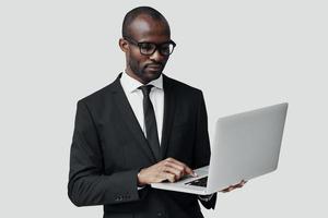 upptagen ung afrikansk man i formell klädsel arbetssätt använder sig av dator medan stående mot grå bakgrund foto